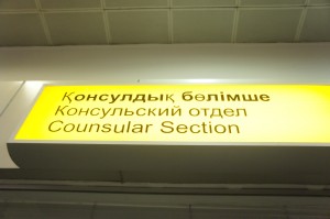 The image shows a sign with text in three languages. The text reads "Қонсулдық бөлімше" in Kazakh, "Консульский отдел" in Russian, and "Counsular Section" in English. The sign is illuminated with a yellow background and black text.