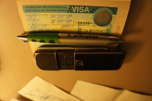 The image shows a Kazakhstan visa document, a pen, and a mobile phone placed on a surface. The visa document is partially visible, with some text and a seal. The pen has the text "BoardingArea" on it, and the mobile phone is a classic model with a physical keypad. There are also some papers partially visible in the background.