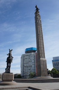 The image shows a tall monument with a statue of a person on top, situated in an urban area. In the foreground, there is a statue of a person on horseback. Behind the monument, there is a modern multi-story building with a blue sign on its roof. The sky is clear with a few clouds.