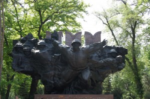 The image shows a large, dark-colored monument featuring multiple human figures in dynamic poses. The central figure appears to be reaching out with an arm extended. The monument is set against a backdrop of green trees and a bright sky. The base of the monument has some text inscribed on it.