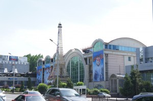 The image shows a large building with a distinctive architectural design, featuring arched windows and a small replica of the Eiffel Tower in front. The building is adorned with large banners and is surrounded by greenery. Several cars are parked or driving in the foreground. The sky is clear and bright.