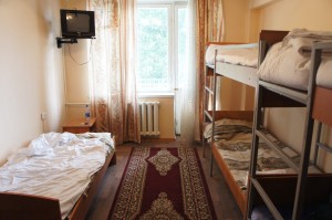 The image shows a small, tidy room with three beds. On the left side, there is a single bed with white sheets. On the right side, there is a bunk bed with the top and bottom bunks made up with bedding. A red and beige patterned rug runs down the center of the room. At the far end of the room, there is a window with sheer curtains, allowing natural light to enter. A small radiator is positioned below the window. A small wooden table with a water bottle on it is placed next to the single bed. A small television is mounted on the wall above the table.