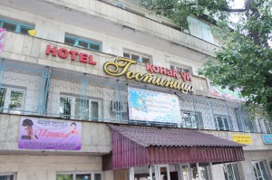 The image shows the exterior of a multi-story building with signs indicating that it is a hotel. The building has a sign in both English and Cyrillic script that reads "HOTEL" and "Гостиница" (which means "hotel" in Russian). There are several banners and signs on the building, including one with a picture of a woman and another with decorative patterns. The building has a metal awning above the entrance and is surrounded by trees.