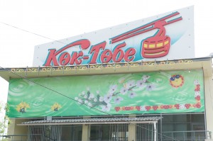 The image shows a building with a large sign on top that reads "Kok-Tobe" in red letters. The sign also features an illustration of a red cable car. Below the sign, there is a green banner with floral designs and a small circular emblem on the right side. The building has a metal railing and several windows.