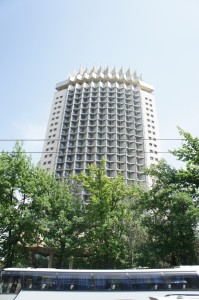 The image shows a tall, modern building with a unique architectural design featuring numerous balconies or protrusions on its facade. The building is surrounded by green trees, and there is a blue sky in the background. The lower part of the image includes a partial view of a bus or vehicle with a blue roof.