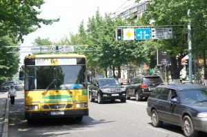 The image shows a busy street with several vehicles, including a yellow and green bus on the left side and multiple cars on the right. The street is lined with trees and there are traffic lights and signs overhead. A pedestrian is walking on the sidewalk to the left of the bus. The scene appears to be in an urban area with buildings visible in the background.