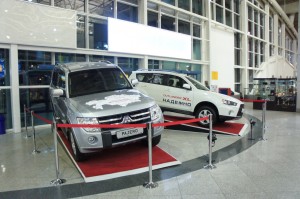 The image shows two cars on display inside a building, possibly an airport or a showroom. The cars are placed on raised platforms with red carpets and are surrounded by stanchions with red ropes. The car on the left is a silver Mitsubishi Pajero, and the car on the right is a white Mitsubishi Outlander XL. The background features large windows and a high ceiling, indicating a spacious and well-lit area.