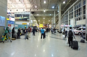 The image shows a busy airport terminal with people walking and standing around. Some individuals are carrying or pulling luggage. There are various signs and advertisements on the walls, and the terminal has a high ceiling with exposed beams and lights. To the right, there are large windows and a row of parked vehicles, possibly taxis or shuttles. The overall atmosphere is bustling, typical of an airport environment.