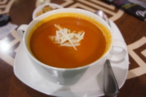 A white bowl filled with tomato soup, garnished with shredded cheese, is placed on a white saucer. A spoon rests on the saucer next to the bowl. The background shows a wooden table with a geometric pattern and some blurred items.