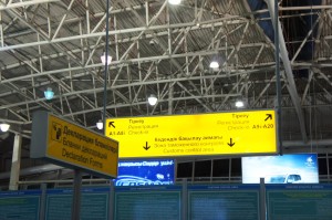 The image shows the interior of an airport terminal with overhead signs. The signs are in multiple languages, including English and Cyrillic script. The yellow sign directs passengers to gates A1-A6 and A11-A20 for check-in and to the customs control area. Another sign indicates the direction to the departure gates. The ceiling of the terminal is visible, with metal beams and lights.