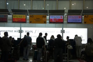 The image shows a group of people standing in line at an airport check-in counter. Above the counters, there are several digital screens displaying flight information and gate numbers. The area is well-lit, and the background includes signage with text in a non-English language. Some people are handling their luggage while waiting in line.