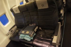 The image shows a row of empty airplane seats with dark leather upholstery. On one of the seats, there is a folded blanket and a small, closed tray table. The seats have headrests with light-colored covers, and the window next to the seats shows a view of the sky. The overall setting appears to be the interior of an airplane.
