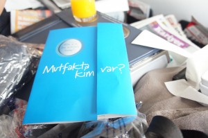 A blue book with the text "Mutfakta Kim var?" on the cover is placed on a cluttered surface. The background includes various items such as a yellow drink, papers, and other miscellaneous objects.