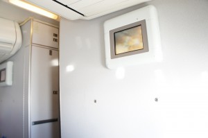 The image shows the interior of an airplane, specifically a section of the wall near the lavatory or galley area. There is a small screen or monitor mounted on the wall, displaying an image that is not clearly discernible. The wall is light-colored, and there are some storage compartments or cabinets visible to the left side of the image. The lighting is bright, suggesting it is daytime.