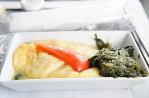 A rectangular white dish contains an omelette topped with a strip of red bell pepper and a serving of cooked spinach on the side. The dish is placed on a white surface, possibly a tray.