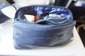 A small, partially open blue toiletry bag is sitting on a white surface. The bag contains various items, including a small bottle and other personal care products. The bag has a logo and text on the front.