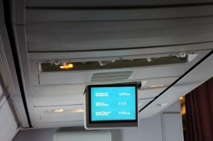 The image shows the interior of an airplane, specifically the overhead panel and a screen. The screen displays flight information, including the distance to the destination, the time at the destination, and the estimated time of arrival. The overhead panel includes air vents and a light.