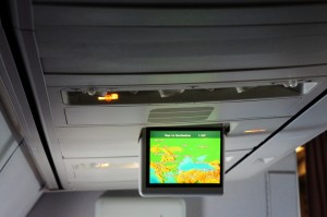 The image shows the interior of an airplane, specifically the overhead panel and a small screen. The screen is displaying a flight map with a green background, indicating the plane's current location and route. The overhead panel includes air vents, reading lights, and a "No Smoking" sign.