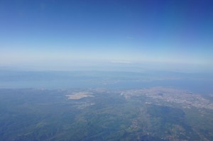 Aerial view of a landscape featuring a mix of green land, urban areas, and a large body of water under a clear blue sky. The horizon is visible in the distance, with a gradient of blue from the sky meeting the land and water below.