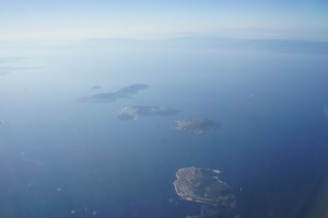Aerial view of several small islands surrounded by a vast expanse of blue water, with a hazy horizon in the background. The islands are scattered and vary in size, with some appearing more developed than others. The sky above is clear with a light blue gradient.