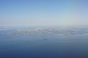 Aerial view of a coastal city with a large body of water in the foreground. The city appears to be densely built with numerous buildings and structures. Several ships or boats are visible in the water near the coast. The sky is clear with a slight haze, and the horizon is visible in the distance.