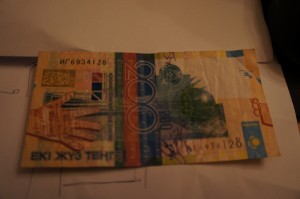 The image shows a 200 Kazakhstani tenge banknote. The banknote features various colors, including yellow, blue, and green, and has intricate designs and patterns. There are also some text and numbers visible on the banknote.