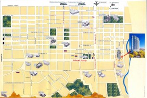 A detailed map of a city area with various buildings and landmarks illustrated. The map includes a grid of streets and avenues, with some labeled, such as "Adams Ave" in red text. There are several notable buildings and structures depicted, including a tall, modern building highlighted on the right side of the map near a river. The map also shows green spaces and parks scattered throughout the city. The top of the map features some transportation icons, possibly indicating train or bus stations. The bottom of the map has a depiction of a mountainous or hilly area.