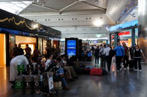The image shows a busy airport terminal with numerous people sitting and standing around. Some individuals are seated on benches, while others are sitting on the floor with their luggage. There are several shops and stores visible in the background, and a large digital display screen is centrally located. The terminal has a high ceiling with modern architectural elements. Recycling bins are also visible in the foreground.