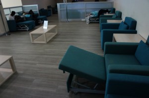 The image shows a waiting area with several teal-colored reclining chairs and wooden side tables. The floor is made of light-colored wood. There are a few people sitting or lying on the chairs in the background. The area appears to be quiet and designed for comfort, possibly in a medical or office setting.