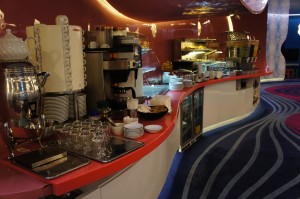 The image shows a modern, well-lit buffet area with a curved counter. The counter is equipped with various kitchen appliances, including a coffee machine, a hot water dispenser, and a toaster. There are stacks of plates, cups, and glasses neatly arranged on the counter. Behind the counter, there are shelves with additional kitchen equipment and food items. The floor has a colorful, wavy-patterned carpet, and the overall ambiance appears to be clean and organized.