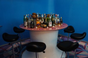 The image shows a round bar table with a pink top, surrounded by five black bar stools. On the table, there is a large assortment of various liquor bottles, including some with recognizable labels. The background is a solid blue wall, and the floor has a colorful, abstract pattern.