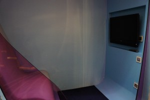 The image shows a small, enclosed room with blue walls. There is a flat-screen television mounted on the right wall. The left side of the image features a translucent, purple-tinted barrier or curtain. The room appears to be dimly lit.