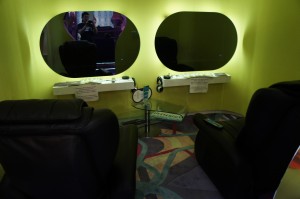 The image shows a room with two black recliner chairs facing a wall with two oval mirrors. The wall is painted a bright yellow-green color. Below each mirror, there is a small white shelf with various items on it, including headphones. There is a glass table between the two chairs, and a remote control is placed on the right chair's armrest. The floor has a colorful, abstract-patterned carpet.