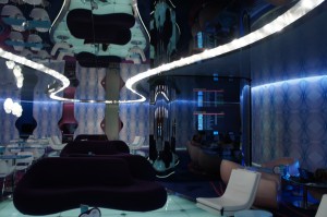 The image shows a modern, futuristic interior space with a unique design. The room features curvy, abstract furniture, including dark purple sofas and white chairs. The ceiling and walls have reflective surfaces, creating a mirrored effect. The lighting is ambient, with blue and white lights illuminating the space, giving it a sleek and contemporary feel. The overall atmosphere is stylish and avant-garde.