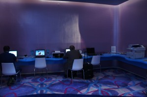 The image shows a dimly lit room with a modern, purple-themed interior. There are two people sitting at a long, blue counter, each working on a laptop. The counter has several other electronic devices, including a desktop computer and a printer. The floor has a colorful, geometric-patterned carpet. The overall ambiance of the room suggests it is a business center or a workspace.