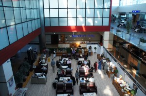 The image shows an indoor area of an airport lounge. The lounge has a modern design with large windows allowing natural light to fill the space. There are several seating areas with people sitting and walking around. Some people are gathered near a counter, possibly for check-in or service. The lounge is furnished with comfortable chairs and tables, and there are plants and other decor elements adding to the ambiance. In the background, there is a sign that reads "Business Lounge." The overall atmosphere appears to be calm and organized.