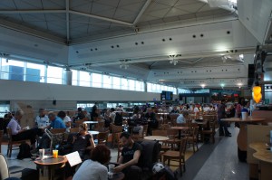 The image shows a busy airport terminal with many people seated at tables, likely in a dining or waiting area. The terminal has a high ceiling with large windows allowing natural light to fill the space. There are various groups of people engaged in different activities such as eating, using laptops, and conversing. The overall atmosphere appears to be bustling and lively.
