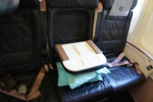The image shows a row of airplane seats. The seats are upholstered in dark material, and the middle seat has a tray table placed on it. There are also some folded blankets or pillows on the middle seat. The seat belts are visible, and the window of the airplane can be seen on the right side of the image.