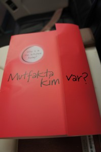 The image shows a red book with the title "Mutfakta Kim var?" written in black text on the cover. The book is placed on a surface, and part of a seat and a seatbelt are visible in the background, suggesting it might be on an airplane or a similar setting.