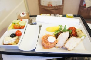 A meal tray with various dishes is shown in the image. The tray includes a main plate with slices of chicken breast, a small portion of smoked salmon with a dollop of cream, a piece of yellow polenta, and some vegetables. There is also a side dish with a small salad containing cherry tomatoes, cucumber slices, and olives. Additionally, there is a dessert in a small glass, possibly a mousse topped with a cherry. The tray also has a rolled-up napkin, a fork, and a knife.