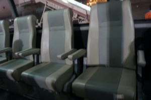 The image shows a row of three empty airplane seats. The seats are upholstered in a combination of light and dark gray fabric, with armrests on either side. The background includes part of the airplane's interior and a glimpse of the outside through the window.