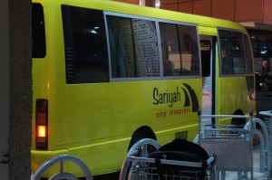 The image shows a yellow bus with the text "Sariyah" and "city express" written on its side. The bus is parked near a building, and there are several luggage carts in the foreground. The scene appears to be at a transportation hub, possibly an airport or a bus station.