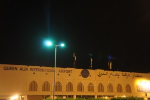 The image shows the exterior of Queen Alia International Airport at night. The building is illuminated by lights, and the name of the airport is displayed in both English and Arabic on the facade. There are several arched windows and a few flags on top of the building. A streetlight is visible in the foreground.