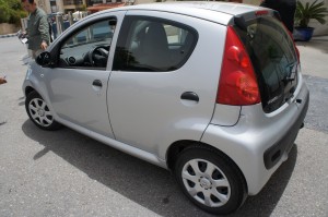 A small silver hatchback car is parked on a street. The car has four doors and is viewed from the rear passenger side. The rear lights are red, and the car has white wheel rims. There are a few people visible in the background.