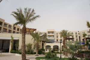 The image shows a modern hotel or resort building with multiple floors and balconies. The architecture is light-colored and surrounded by a well-maintained garden area featuring several palm trees and other greenery. The sky appears overcast, and there are pathways and outdoor seating areas visible in the foreground.