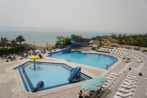 The image shows an outdoor swimming pool area with two pools. The larger pool has a blue water slide and is surrounded by numerous white lounge chairs. The smaller pool has a yellow mushroom-shaped water feature and a blue dolphin statue. There are a few people sitting under green and white striped umbrellas near the pools. In the background, there are palm trees and a body of water, possibly a sea or lake, under a hazy sky.