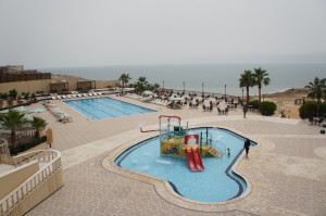 The image shows an outdoor swimming pool area at a resort by the sea. There are two pools: a larger rectangular pool and a smaller, irregularly shaped children's pool with a water slide and play structure. The area is surrounded by lounge chairs and palm trees, with the beach and ocean visible in the background. The sky appears overcast.