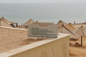 The image shows a beach scene with several thatched-roof umbrellas in the background near the shoreline. In the foreground, there is a sign with instructions for swimmers at the Dead Sea. The sign reads:
- Do not dive.
- Do not swim far from shore.
- Protect eyes and mouth from water.
- Try to swim on your back.
- Remember there is no lifeguard on duty.