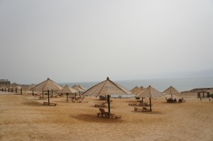 The image shows a beach with several thatched-roof umbrellas and wooden lounge chairs arranged in rows on the sandy shore. The sea is visible in the background under a hazy sky. The beach appears to be relatively empty, with no people in sight.