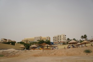 The image shows a beach scene with several thatched umbrellas and lounge chairs on the sandy shore. In the background, there are multiple multi-story buildings, likely hotels or resorts, surrounded by greenery and palm trees. The sky is overcast, giving the scene a muted, calm atmosphere.